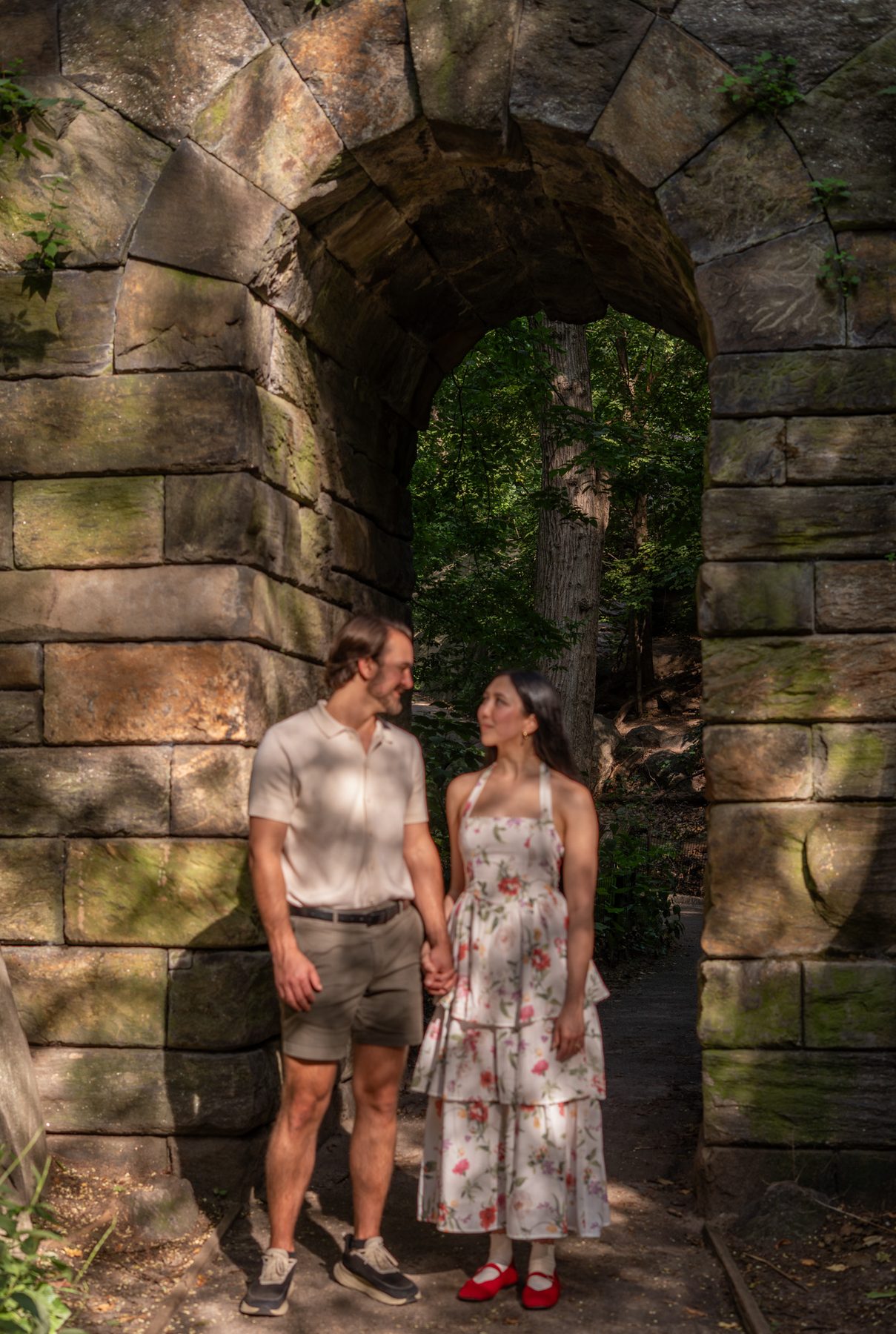 A couple holding hands under a stone archway in dappled summer light