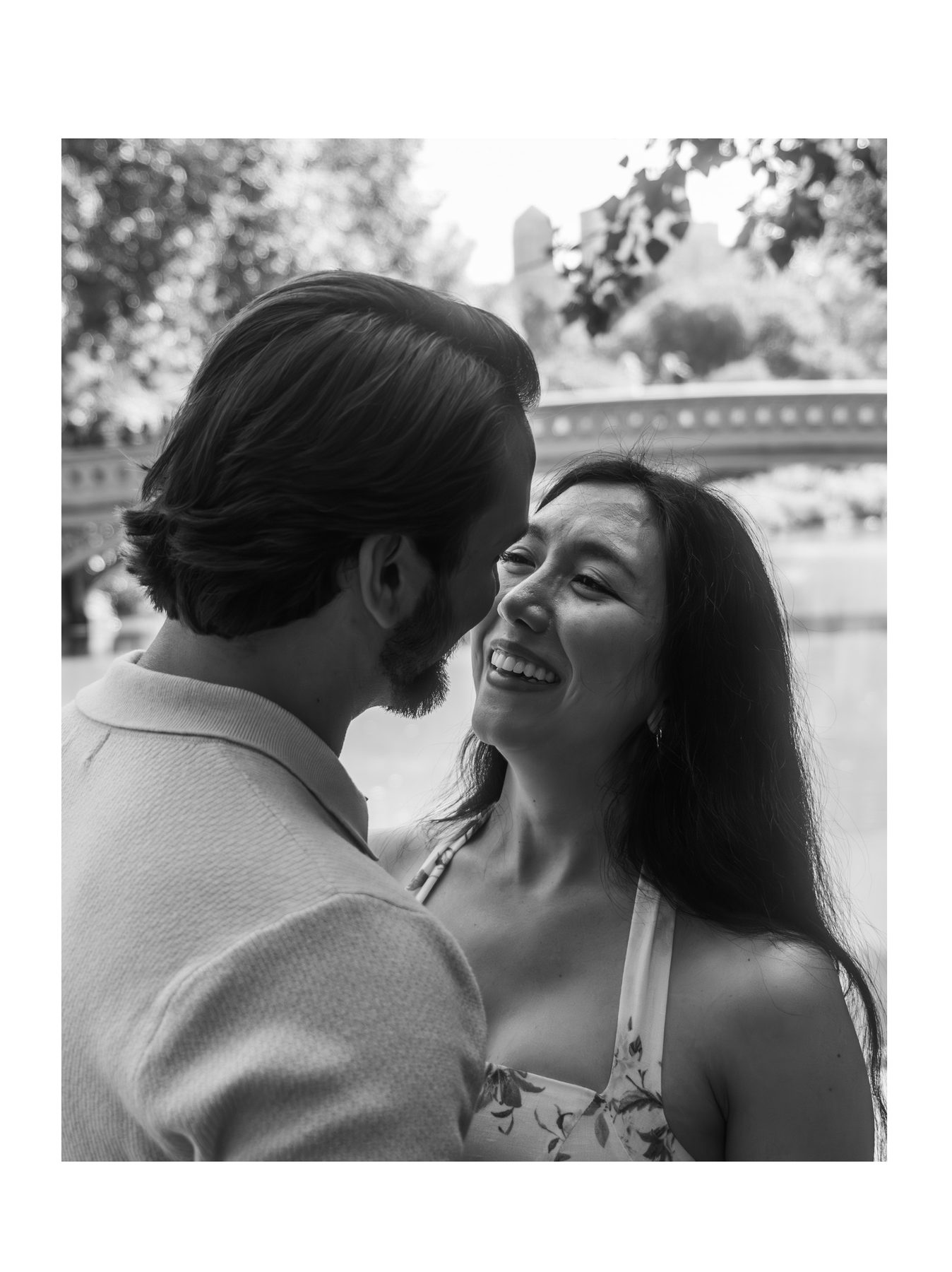 A black and white portrait of a couple, she laughs as he leans in, a park bridge behind