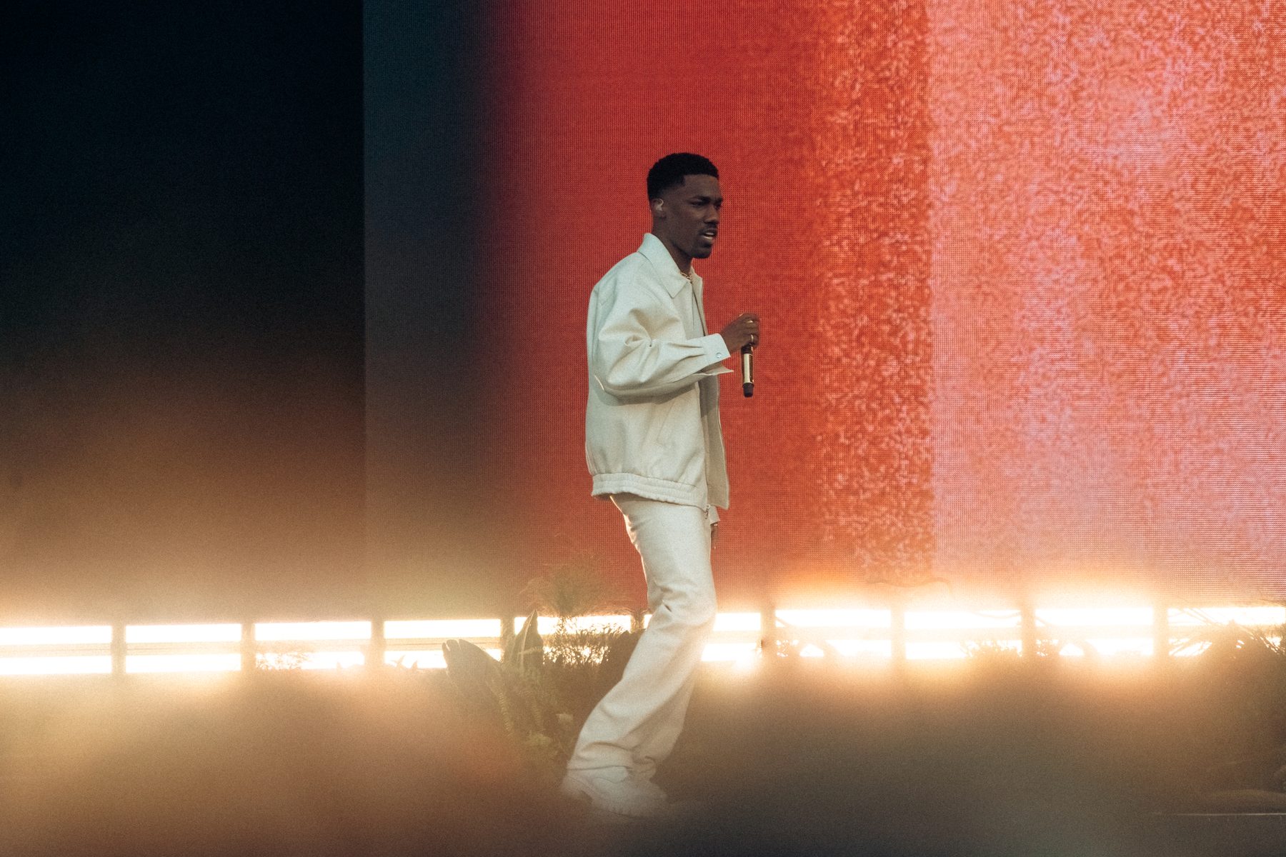 A performer in a white suit crossing a wide stage, backlit by a red LED wall