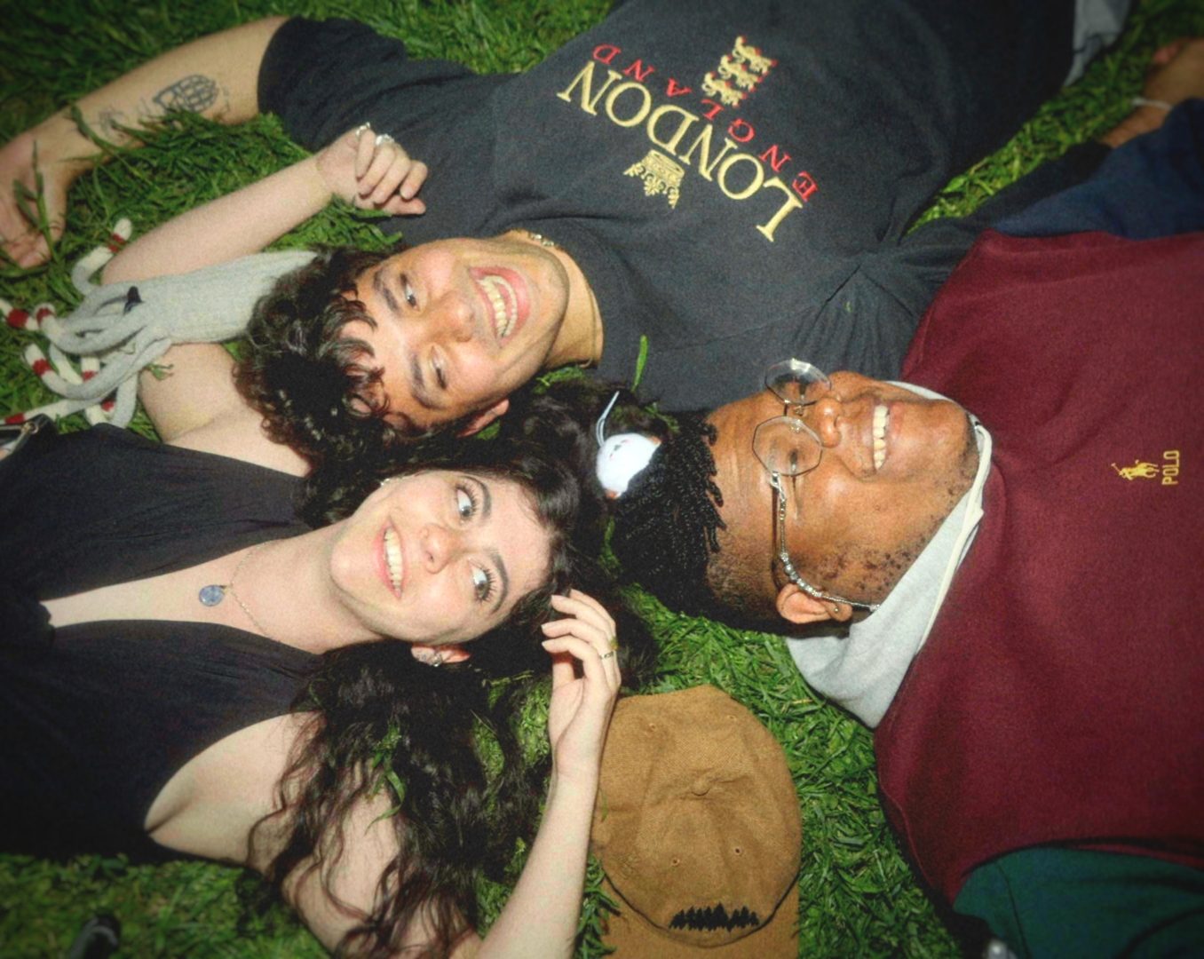 Three friends lying head to head on grass at night, laughing up at the camera